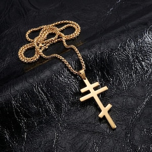 May include: Gold-colored necklace featuring a double-barred cross pendant. The pendant hangs from a matching chain, displayed against a dark, textured background. The cross design is a prominent religious symbol.