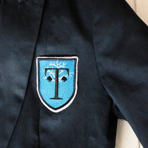 May include: A black blazer with a blue and white embroidered patch featuring a tree and the letter "T".