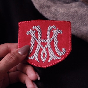 Puede incluir: Parche rojo bordado con forma de escudo. Presenta un diseño de monograma blanco estilizado. Los bordes están cuidadosamente cosidos con hilo rojo. El parche se muestra sobre un fondo oscuro.