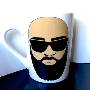 Puede incluir: Taza de cerámica blanca con un gráfico de un hombre calvo con gafas de sol y una barba negra completa. La taza tiene un asa curva y un acabado brillante. El diseño está centrado en la parte delantera de la taza.