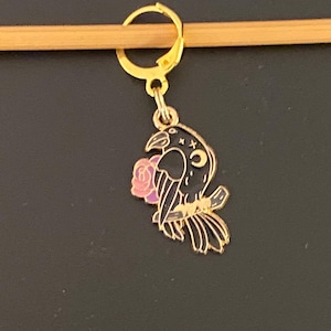 May include: A gold-colored metal stitch marker with a black raven design holding a pink rose. The raven has a crescent moon and star detail. The stitch marker is attached to a gold-colored hoop.