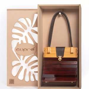 May include: A wooden handbag with a dark brown leather strap and a gold clasp, presented in a wooden box. The bag features a combination of light and dark wood tones. The box includes the brand name "cupre" and a leaf design.