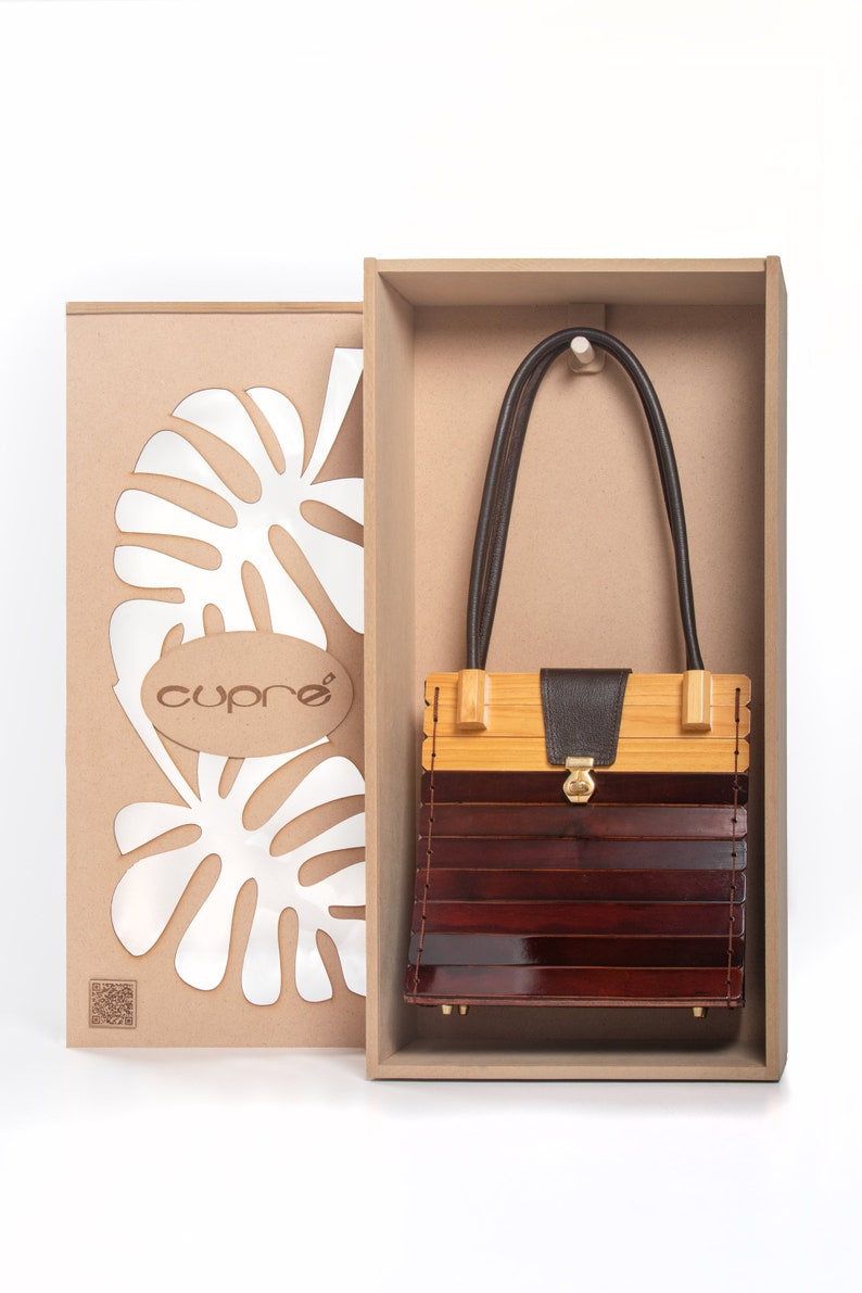 May include: A wooden handbag with a dark brown leather strap and a gold clasp, presented in a wooden box. The bag features a combination of light and dark wood tones. The box includes the brand name "cupre" and a leaf design.