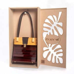 May include: A wooden handbag with a bamboo top and a dark brown, glossy body. The bag has a leather handle and a gold-coloured clasp. It is presented in a cardboard box with a leaf design and the brand name "cupro".