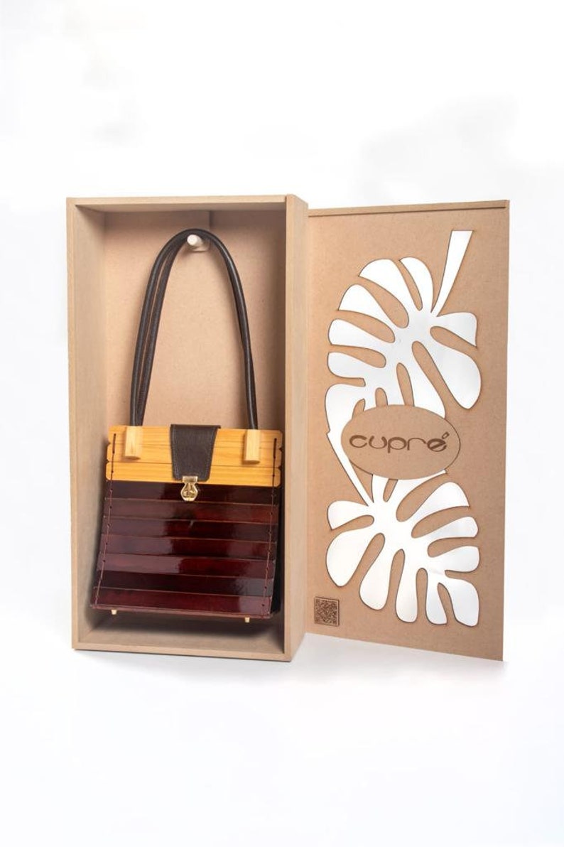 May include: A wooden handbag with a bamboo top and a dark brown, glossy body. The bag has a leather handle and a gold-coloured clasp. It is presented in a cardboard box with a leaf design and the brand name "cupro".