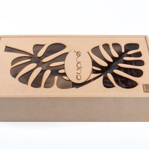 May include: A rectangular wooden box with a lid featuring leaf-shaped cutouts. The box is light brown and has the word "Cupr&eacute;" printed on an oval-shaped piece of wood. The box is likely a gift box or storage container.
