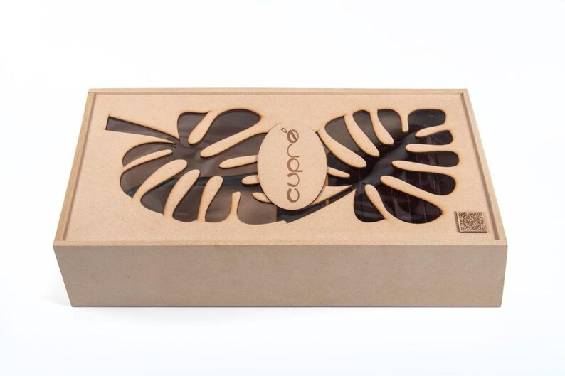 May include: A rectangular wooden box with a lid featuring leaf-shaped cutouts. The box is light brown and has the word "Cupr&eacute;" printed on an oval-shaped piece of wood. The box is likely a gift box or storage container.
