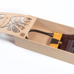May include: A wooden box with a laser-cut lid featuring a monstera leaf design. Inside, a brown and tan handbag with a gold clasp and a black strap rests. The box is open, revealing the bag.