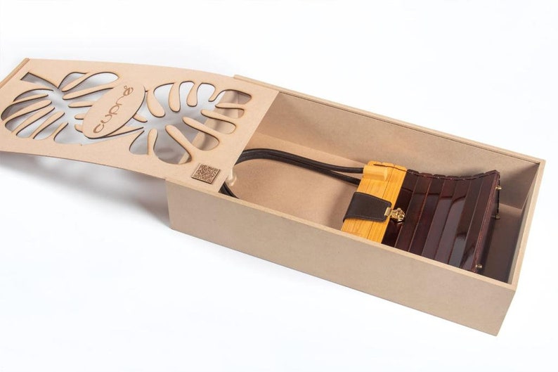 May include: A wooden box with a laser-cut lid featuring a monstera leaf design. Inside, a brown and tan handbag with a gold clasp and a black strap rests. The box is open, revealing the bag.