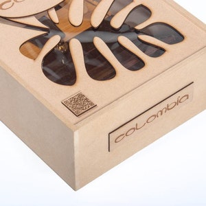 May include: A light brown wooden box with a laser-cut leaf design on the lid. The box has the word "Colombia" on a small rectangular wooden plaque. A QR code is also visible.