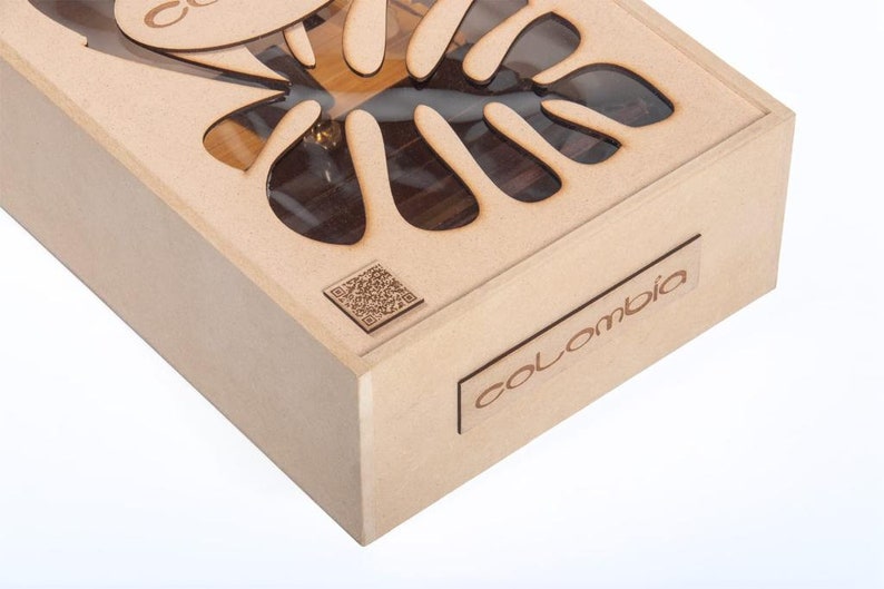 May include: A light brown wooden box with a laser-cut leaf design on the lid. The box has the word "Colombia" on a small rectangular wooden plaque. A QR code is also visible.