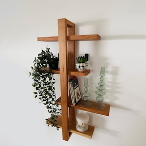 May include: A wooden wall shelf with five shelves. The shelves are decorated with a potted plant, a small potted succulent, a book, two glass candle holders, and a white candle.