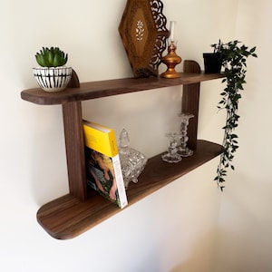 K&ouml;nnte beinhalten: Ein zweistufiges Wandregal aus Holz mit einem kleinen Topf mit einer Sukkulente, einem dekorativen Tablett, einem Kerzenhalter und einem Buch. Das Regal ist an einer wei&szlig;en Wand montiert.