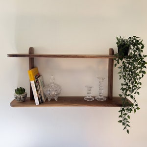 K&ouml;nnte beinhalten: Ein Holzregal mit zwei Ablagen, einem kleinen Sukkulenten im Topf, einem Glasgef&auml;&szlig; mit Deckel, zwei Glaskerzenhaltern und einem Stapel B&uuml;cher. Eine H&auml;ngepflanze h&auml;ngt von der oberen Ablage.