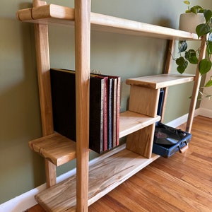 May include: A light-colored wooden record shelf with multiple shelves and vertical supports. The shelf holds vinyl records and a small record player. A potted plant sits on the top shelf. The shelf is made of natural wood.