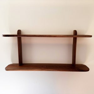 K&ouml;nnte beinhalten: Ein zweistufiges Holzregal aus Walnussholz. Das Regal ist an einer wei&szlig;en Wand montiert.