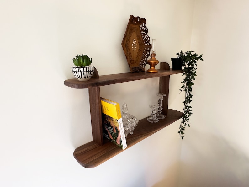 K&ouml;nnte beinhalten: Ein zweistufiges Wandregal aus Holz mit einem kleinen Topf mit einer Sukkulente, einem dekorativen Tablett, einem Kerzenhalter und einem Buch. Das Regal ist an einer wei&szlig;en Wand montiert.