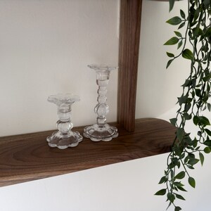 K&ouml;nnte beinhalten: Zwei klare Glaskerzenhalter mit einem dekorativen, strukturierten Design stehen auf einem Holzregal. Das Regal ist Teil einer Wandmontage mit einer dunklen Holzoptik.