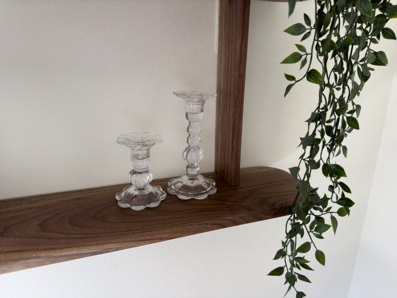 K&ouml;nnte beinhalten: Zwei klare Glaskerzenhalter mit einem dekorativen, strukturierten Design stehen auf einem Holzregal. Das Regal ist Teil einer Wandmontage mit einer dunklen Holzoptik.