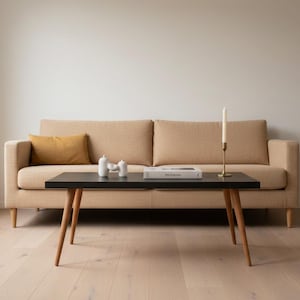 May include: A beige sofa with a mustard cushion and a black coffee table with wooden legs. The table holds white ornaments, a book, and a gold candle holder with a lit candle. The room has light-coloured wood flooring.