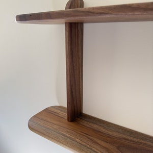 K&ouml;nnte beinhalten: Ein zweistufiges Holzregal mit abgerundeten Kanten aus Walnussholz mit nat&uuml;rlicher Oberfl&auml;che. Das Regal ist an einer Wand montiert und hat ein schlichtes, minimalistisches Design.