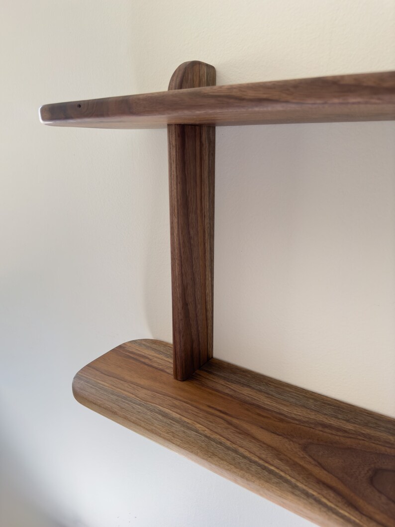 K&ouml;nnte beinhalten: Ein zweistufiges Holzregal mit abgerundeten Kanten aus Walnussholz mit nat&uuml;rlicher Oberfl&auml;che. Das Regal ist an einer Wand montiert und hat ein schlichtes, minimalistisches Design.
