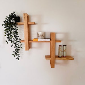 May include: A wooden wall shelf with a geometric design. The shelf has three levels and is decorated with a potted plant, a candle, a book titled "plant magic", and two rolled-up items.
