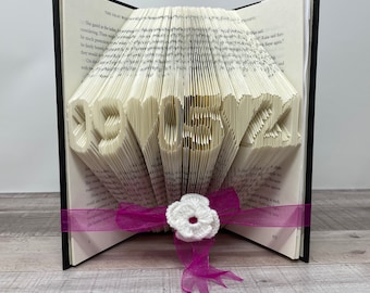 May include: A book with the pages folded into the shape of the numbers 1952. The book is open and a pink ribbon with a white crocheted flower is tied around it.
