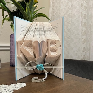 May include: A folded book art sculpture featuring the letters K and B separated by a central heart shape. The pages are intricately creased to form these characters. A sheer white ribbon is tied around the book with a small turquoise flower.