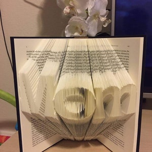 May include: A book with the word "LOVE" folded into the pages. The book is open and the pages are folded to create the word. The book is on a wooden table.