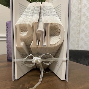 May include: A handmade book art piece with the word "DAD" folded into the pages. The book is tied with a white ribbon and bow. The pages are light beige, and the cover is gray.