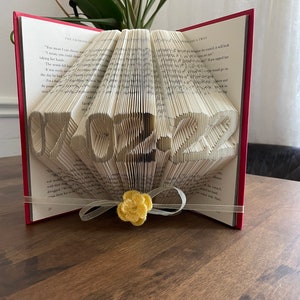 May include: A book with pages folded into the shape of the year 2022. The book is red and has a white ribbon tied around it with a yellow flower.