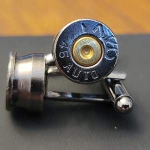 May include: A pair of cufflinks made from .45 Auto shell casings. The casings are silver with black lettering and a gold-colored primer. The cufflinks are attached to a silver metal bar.