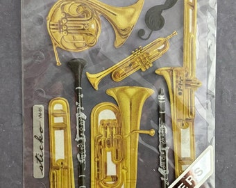 May include: A package of vellum stickers featuring various gold-colored musical instruments, including a trumpet, trombone, tuba, clarinet, and French horn. The package also includes a flute and a musical note. The text "Vellum Stickers" is visible.