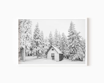 Könnte beinhalten: Ein Schwarzweißfoto einer kleinen weißen Hütte mit einer Holztür und einem weißen Lattenzaun. Die Hütte ist von schneebedeckten Kiefern umgeben.