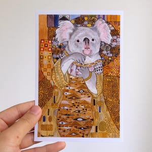 Puede incluir: Una postal con un koala en una túnica dorada, inspirada en el arte de Gustav Klimt. El koala, con pelaje gris, es el centro, sobre un fondo detallado de patrones dorados y marrones. La obra de arte está sujeta por una mano.