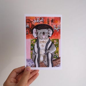 Puede incluir: Una impresión en acuarela que presenta un koala con un sombrero negro y un chal a rayas. El koala es la figura central, sobre un fondo de una escena rosa y roja con elementos florales y arquitectónicos. La obra de arte tiene un estilo ilustrativo caprichoso.