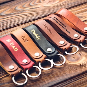 Könnte beinhalten: Eine Sammlung personalisierter Leder-Schlüsselanhänger in verschiedenen Farben, darunter Braun, Rot und Schwarz. Jeder Schlüsselanhänger hat einen silbernen Schlüsselring und ist mit Namen oder Phrasen wie "#1 DAD" und "George" graviert. Die Schlüsselanhänger liegen auf einer Holzoberfläche.