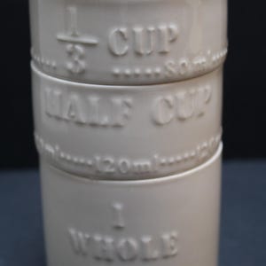 May include: A stack of three off-white ceramic measuring cups. The cups are labeled with embossed text: 1/3 CUP, HALF CUP, and 1 WHOLE CUP. The 1/3 cup is 80ml, and the half cup is 120ml. The cups have a simple, classic design.
