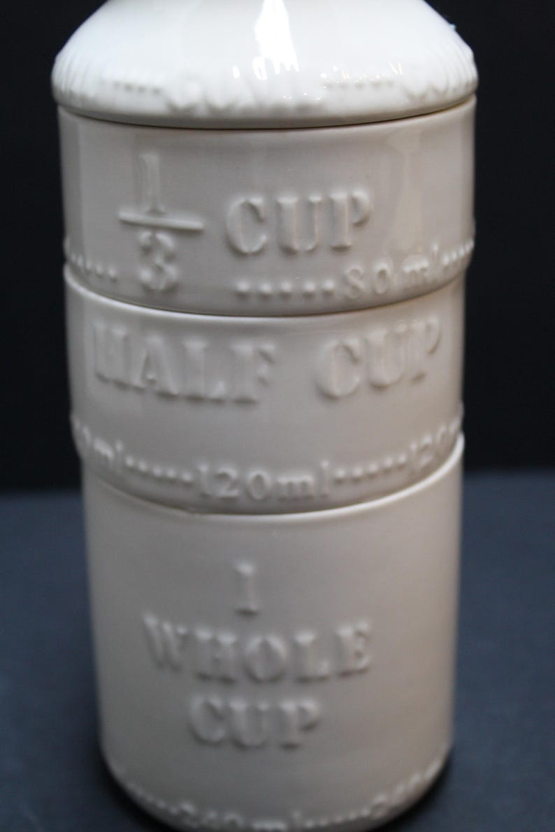 May include: A stack of three off-white ceramic measuring cups. The cups are labeled with embossed text: 1/3 CUP, HALF CUP, and 1 WHOLE CUP. The 1/3 cup is 80ml, and the half cup is 120ml. The cups have a simple, classic design.