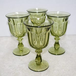 May include: Four green-tinted glass goblets with a tulip-shaped bowl and a decorative stem. The goblets are arranged in a group, showcasing their elegant design. The glasses are made of a translucent material.