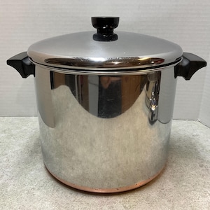 May include: A large stainless steel stockpot with a polished reflective finish, featuring two black side handles and a matching lid with a black knob. The base of the pot shows a distinct copper-colored band around the bottom edge.