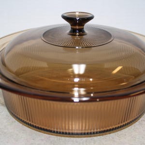 May include: A brown glass casserole dish with a matching lid. The dish has a ribbed design on the sides and lid. The lid has a knob handle. The dish is round and appears to be made of heat-resistant glass.