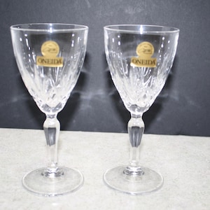 May include: Two clear crystal wine glasses with a decorative cut pattern. Each glass has a stem and a round base. The glasses have a gold label that reads "ONEIDA". The glasses are on a light gray surface.