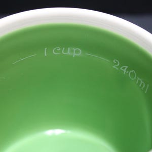 May include: Close-up of a white and green measuring cup. The interior is a vibrant green, with a white rim. The text "1 cup" and "240ml" are etched inside the cup, indicating volume measurements.