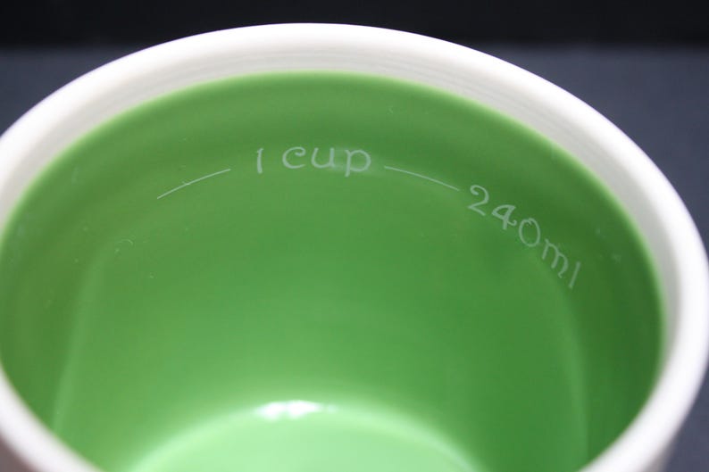 May include: Close-up of a white and green measuring cup. The interior is a vibrant green, with a white rim. The text "1 cup" and "240ml" are etched inside the cup, indicating volume measurements.