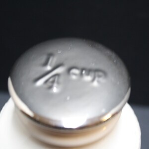 May include: Close-up of a silver measuring cup lid with the text "1/4 cup" embossed on the top. The lid is on top of a white container, set against a dark background.