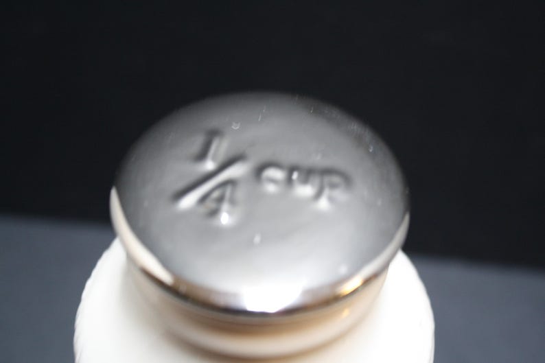 May include: Close-up of a silver measuring cup lid with the text "1/4 cup" embossed on the top. The lid is on top of a white container, set against a dark background.