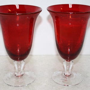 May include: Two ruby red glass goblets with clear stems and bases. The goblets have a flared top and a textured appearance. The glasses are likely used for serving beverages.