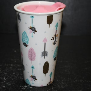 May include: White ceramic travel mug with a pink lid. The mug has a pattern of teal, pink, and brown arrows and leaves.